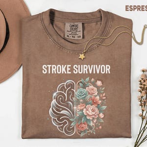 May include: Espresso-coloured t-shirt with the words "STROKE SURVIVOR" printed on the front. The design features a brain graphic and floral elements in pink, blue, and green. A gold necklace with a star pendant is draped across the shirt.