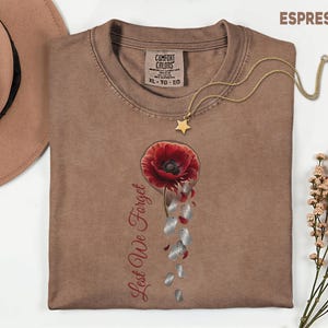May include: Espresso-colored t-shirt with a red poppy graphic and the text "Lest We Forget." A gold star pendant necklace rests on the shirt. The shirt is from Comfort Colors, size XL-TG-EG.