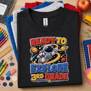 May include: A black short-sleeved t-shirt featuring a graphic of an astronaut floating in space surrounded by planets and stars. The text reads READY TO EXPLORE 3RD GRADE in bold, colorful lettering. The shirt is layered over blue, gray, and red shirts.