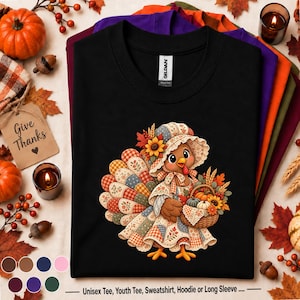 Op de afbeelding: Een zwart t-shirt met een afbeelding van een cartoonkalkoen die een mutsje en een patchworkjurk draagt, met een mandje vol pompoenen en herfstbladeren. De kalkoen heeft een kleurrijke staart met een ruitjes- en bloemenpatroon.