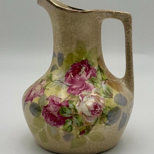 May include: A vintage ceramic pitcher with a beige speckled finish and a gold rim. The pitcher is adorned with a floral design featuring pink and white roses with green leaves. The handle is rectangular.