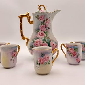 May include: A vintage porcelain tea set featuring a tall, ornate coffee pot and six matching cups. The set is painted with pink roses and gold accents. The coffee pot is approximately 10 inches tall.