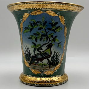 May include: A tall, green vase with a gold rim and base. The vase features a painted scene of a bird perched under a tree, framed by a gold leaf design. The background is a light blue sky.