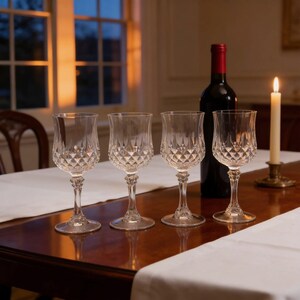 May include: Four clear glass wine goblets featuring a faceted diamond-cut pattern on the lower bowl and a decorative stem. The glasses are arranged in a row next to a dark wine bottle and a single tall white candle in a brass holder.