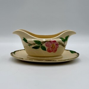 May include: Cream-colored gravy boat and saucer set. The boat features a floral design with pink flowers and green leaves. The set has a gold trim around the edges, adding a touch of elegance. A classic addition to any dining table.