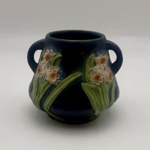 May include: A dark blue ceramic vase with two handles. The vase features a raised floral design with white and orange flowers and green leaves. The vase has a rounded shape and a wide opening.