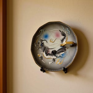 May include: A decorative plate featuring a dragon design in black, white, and gold. The plate has a gold rim and a gold handle. It is mounted on a black stand against a beige wall.