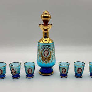 May include: A vintage blue and gold decanter set with six matching shot glasses. The set features a gold-colored stopper and accents, with a cameo design on the decanter and glasses. The set is likely made of glass.