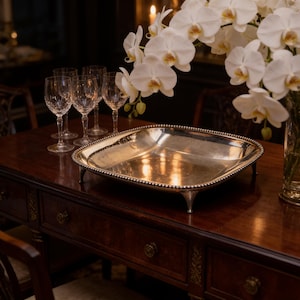 May include: A square silver serving tray with a beaded rim and four short feet. The tray has a polished, reflective surface and is positioned on a dark wooden table alongside several clear glass goblets and a vase of white orchid flowers.