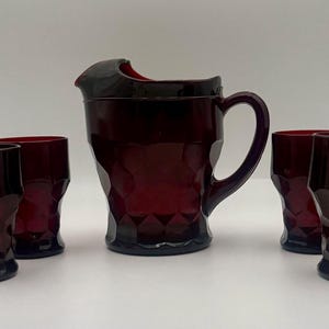 May include: A ruby red glass pitcher and four matching glasses. The pitcher has a handle and a spout. The glasses and pitcher have a textured, geometric design. The set is likely for serving beverages.
