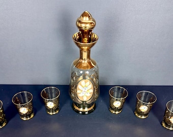May include: A vintage glass decanter set with a gold finish. The set includes a decanter with a stopper and six matching shot glasses. The decanter and glasses feature ornate floral designs.