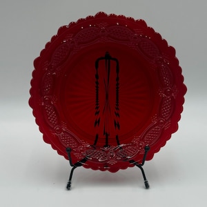 May include: A vibrant red glass plate with a scalloped edge and intricate embossed detailing. The plate is displayed on a black metal stand, showcasing its ornate design. The plate's radiant color and detailed pattern make it a decorative piece.