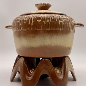 May include: A vintage brown ceramic fondue pot with a lid and handles, resting on a matching wavy-legged stand. The pot has a textured design around the top and a two-tone color scheme of brown and beige.