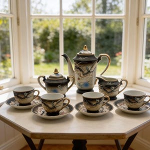 May include: A vintage tea set with a black, gold, and cream dragon design. The set includes a teapot, sugar bowl, creamer, and six teacups with saucers. The tea set is displayed on a small, white table in front of a window.