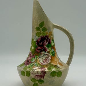 May include: A beige ceramic pitcher with a curved spout and handle. The pitcher is decorated with hand-painted roses in shades of red, pink, and white, surrounded by green leaves. The pitcher has a textured surface and a gold-colored rim.