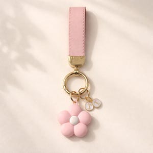 Op de afbeelding: Een roze leren sleutelhanger met een goudkleurige sluiting en ring. Een roze bloemvormige bedel met een wit centrum en twee kleine ronde bedels met de letter "T" hangen aan de ring. Een modieus accessoire.