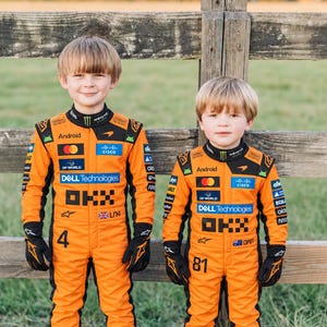 May include: Two children wearing bright orange racing jumpsuits with black accents. The suits feature various sponsor logos including Android, Dell Technologies, and OKX. One suit displays the number 4 and LNA, while the other shows 81 and OP81.