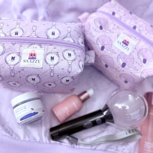 May include: Two lavender cosmetic bags with a zipper closure and a pattern of white graphics. The bags are next to skincare products, a black and clear light stick, and a bottle of perfume. The bags have the text "SNAZZY TOTE".