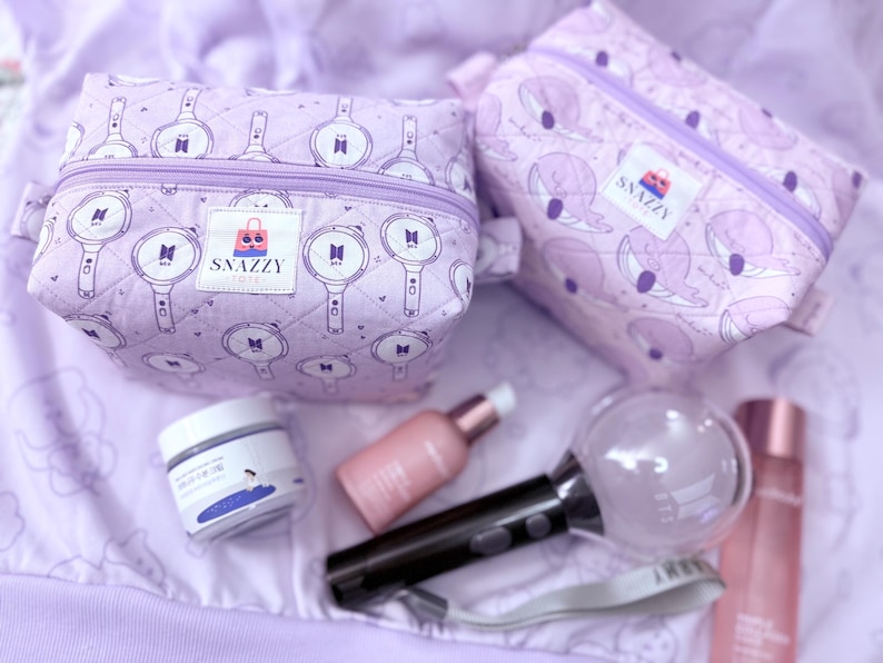 May include: Two lavender cosmetic bags with a zipper closure and a pattern of white graphics. The bags are next to skincare products, a black and clear light stick, and a bottle of perfume. The bags have the text "SNAZZY TOTE".