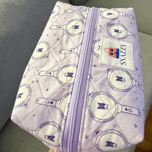 May include: A quilted lavender tote bag with a zipper closure and a matching strap. The bag features a repeating pattern of white and purple graphics. A small label reads "SNAZZY TOTE".