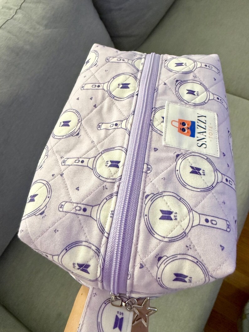 May include: A quilted lavender tote bag with a zipper closure and a matching strap. The bag features a repeating pattern of white and purple graphics. A small label reads "SNAZZY TOTE".