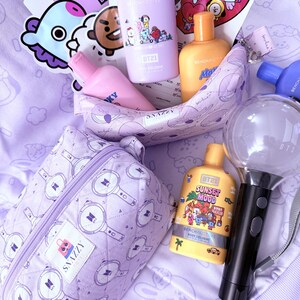 May include: A collection of purple and yellow BT21-themed merchandise, including body cologne bottles, a cosmetic bag, and a light stick. The items are arranged on a light purple surface, with stickers and a heart-shaped card featuring cartoon characters.