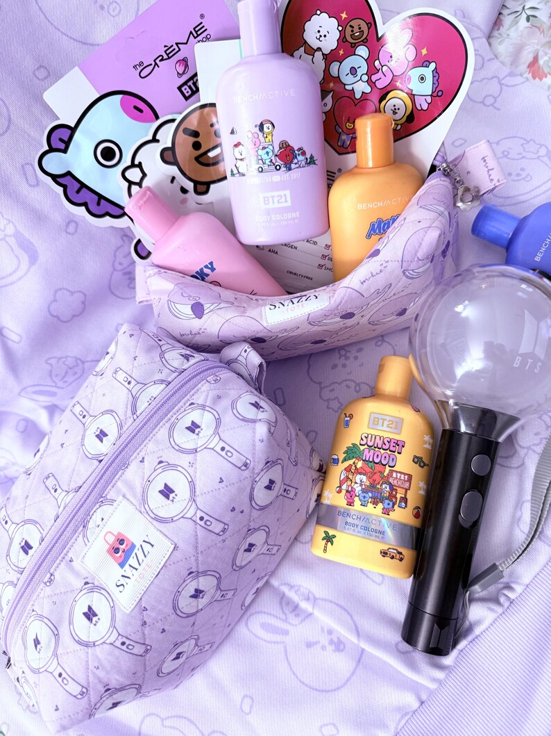 May include: A collection of purple and yellow BT21-themed merchandise, including body cologne bottles, a cosmetic bag, and a light stick. The items are arranged on a light purple surface, with stickers and a heart-shaped card featuring cartoon characters.