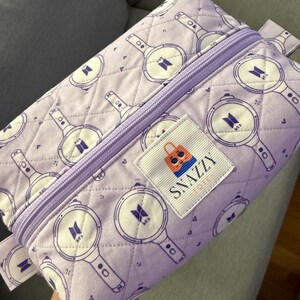 May include: A lavender quilted pencil case with a repeating pattern of purple and white light sticks. The case has a lavender zipper and a label that says "SNAZZY TOTE".