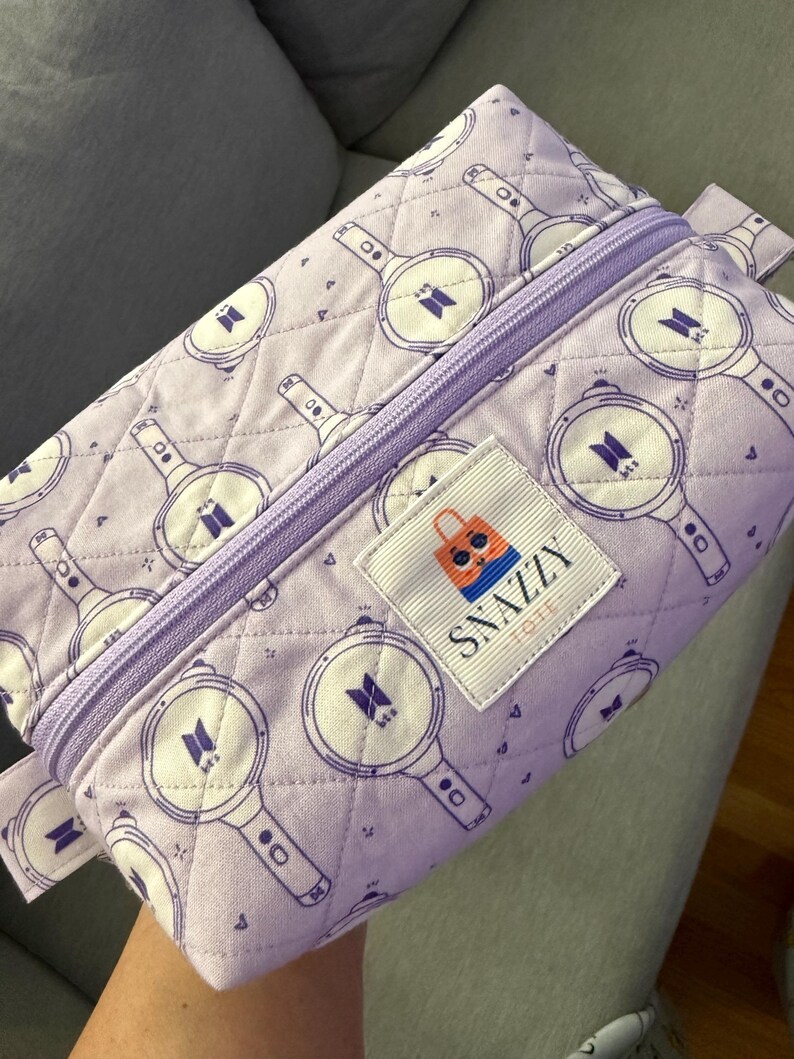 May include: A lavender quilted pencil case with a repeating pattern of purple and white light sticks. The case has a lavender zipper and a label that says "SNAZZY TOTE".