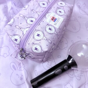 May include: A lavender quilted cosmetic bag with a zipper closure and a star charm. The bag features a repeating pattern of a light stick design. A black and clear light stick is next to the bag. The bag has a label that says "SNAZZY".
