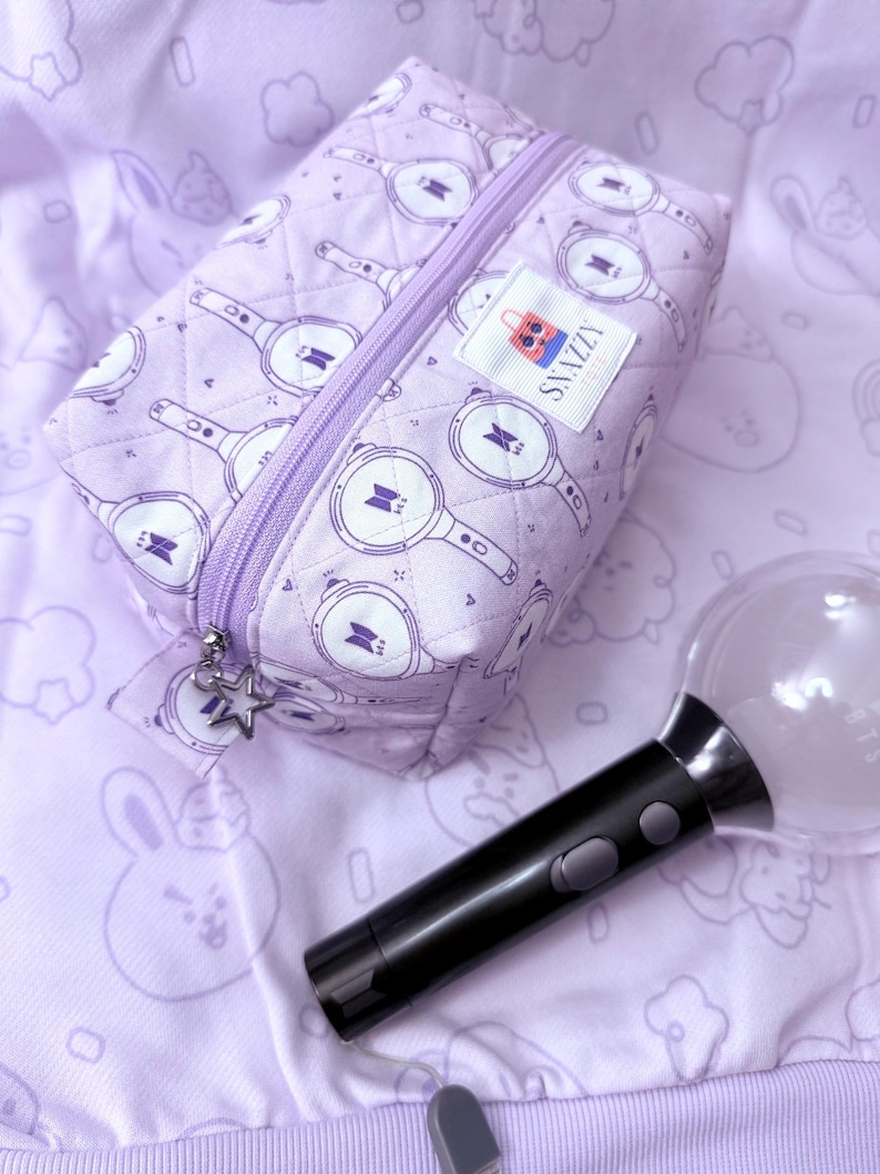 May include: A lavender quilted cosmetic bag with a zipper closure and a star charm. The bag features a repeating pattern of a light stick design. A black and clear light stick is next to the bag. The bag has a label that says "SNAZZY".