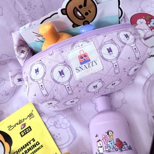 May include: A light purple quilted cosmetic bag with a zipper, featuring a repeating pattern of a microphone and the BTS logo. The bag is open, revealing skincare products and stickers. A bottle of BT21 body cologne is also visible.