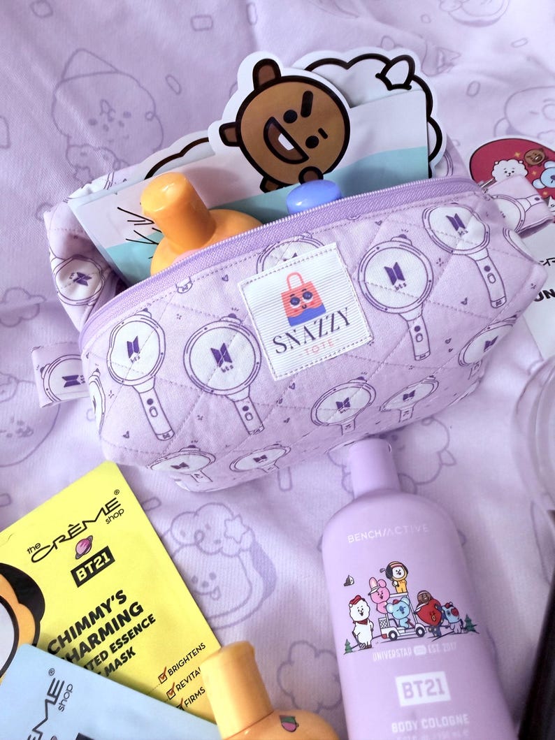 May include: A light purple quilted cosmetic bag with a zipper, featuring a repeating pattern of a microphone and the BTS logo. The bag is open, revealing skincare products and stickers. A bottle of BT21 body cologne is also visible.