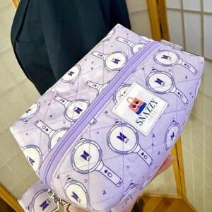 May include: A lavender quilted cosmetic bag with a repeating pattern of white and purple graphics. The bag has a purple zipper, a small fabric tag that says "Snazzy Tote", and a star-shaped charm.
