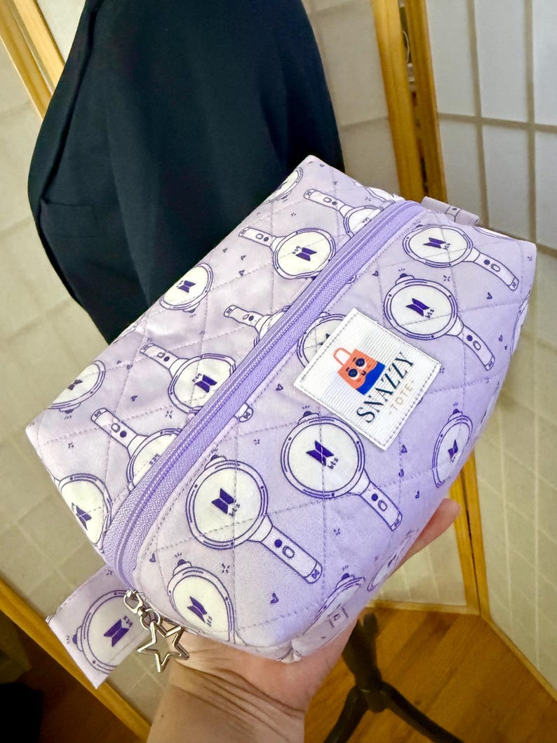 May include: A lavender quilted cosmetic bag with a repeating pattern of white and purple graphics. The bag has a purple zipper, a small fabric tag that says "Snazzy Tote", and a star-shaped charm.