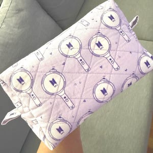 May include: A quilted lavender pouch featuring a repeating pattern of purple magnifying glass designs with a logo in the center. The pouch has a small loop on one side and is likely used for storage or carrying small items.