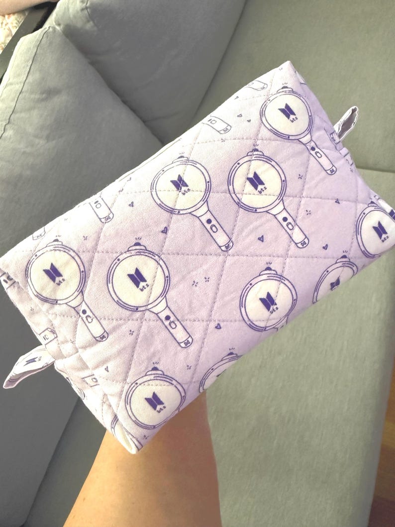 May include: A quilted lavender pouch featuring a repeating pattern of purple magnifying glass designs with a logo in the center. The pouch has a small loop on one side and is likely used for storage or carrying small items.