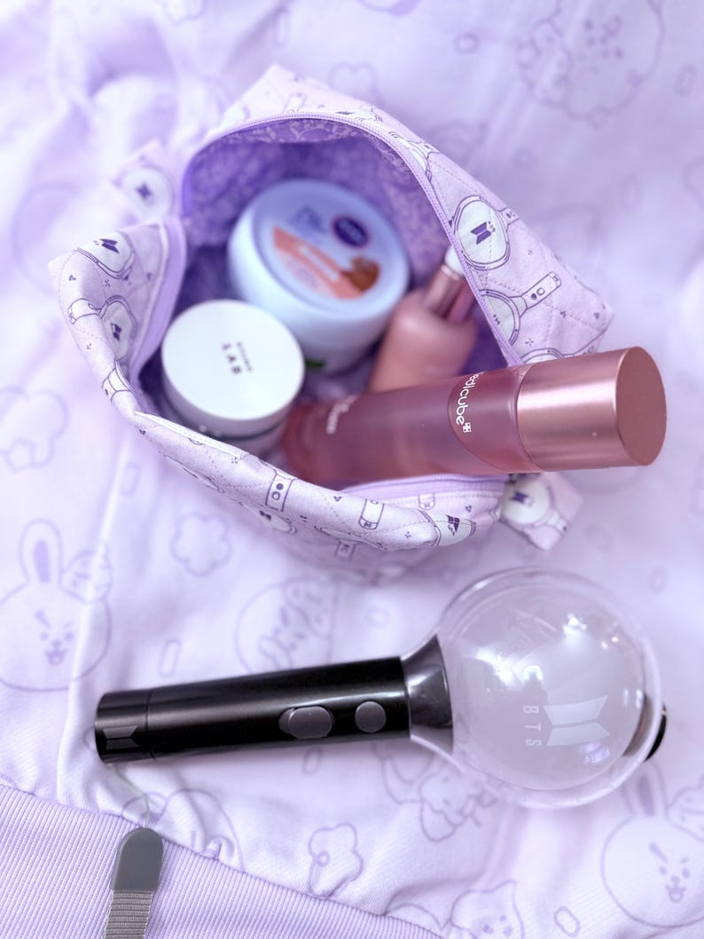 May include: A purple cosmetic bag with a zipper, filled with skincare products. The bag is patterned with a repeating design. A black and clear light stick with the letters "BTS" is next to the bag. The background is a light purple fabric.