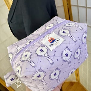 May include: A quilted, lavender cosmetic bag with a repeating pattern of white and purple light sticks. The bag has a lavender zipper and a small tag that says "SNAZZY TOTE". A silver star charm hangs from the zipper pull.