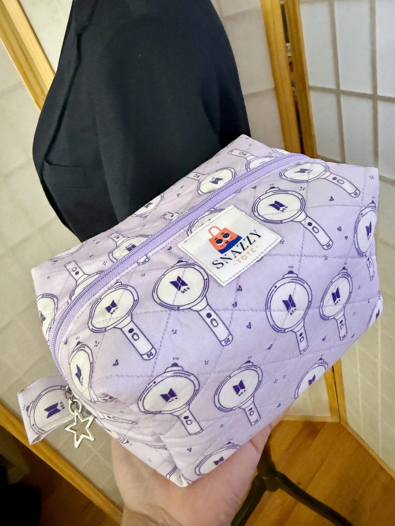 May include: A quilted, lavender cosmetic bag with a repeating pattern of white and purple light sticks. The bag has a lavender zipper and a small tag that says "SNAZZY TOTE". A silver star charm hangs from the zipper pull.