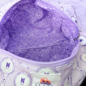 May include: A quilted lavender tote bag with a floral interior lining. The bag features a zipper closure and a label that reads "SNAZZY TOTE". The exterior has a repeating pattern of a logo and a light stick design.