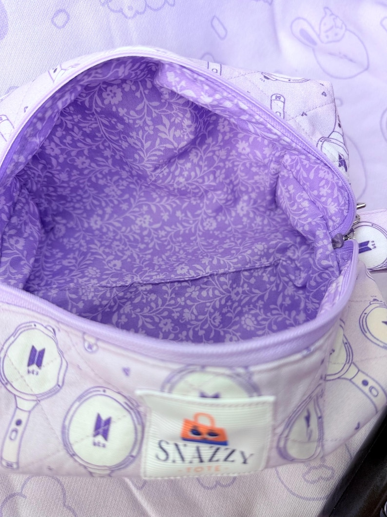 May include: A quilted lavender tote bag with a floral interior lining. The bag features a zipper closure and a label that reads "SNAZZY TOTE". The exterior has a repeating pattern of a logo and a light stick design.