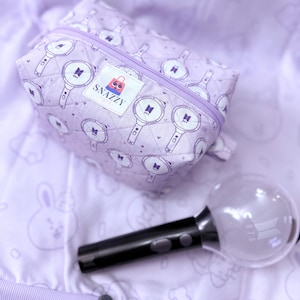 May include: A lavender quilted cosmetic bag with a zipper closure, featuring a repeating pattern of light sticks and the BTS logo. A black light stick with a clear globe and the BTS logo is next to the bag. The bag has a "Snazzy Totes" label.