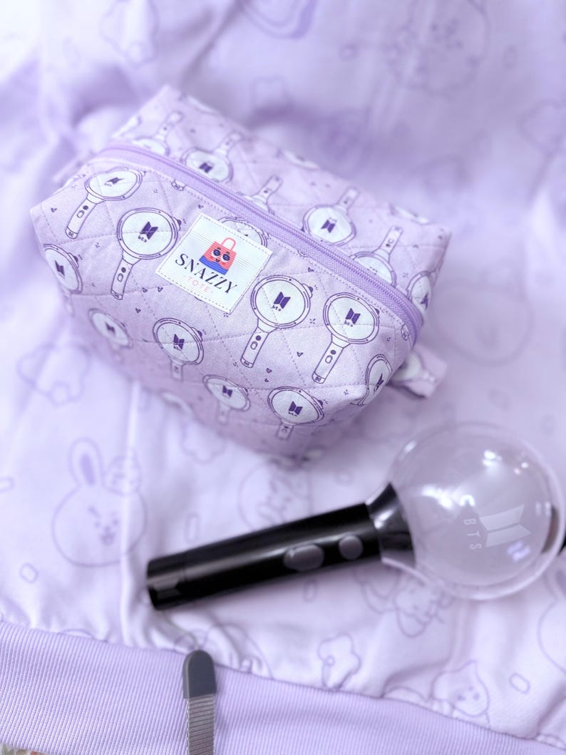 May include: A lavender quilted cosmetic bag with a zipper closure, featuring a repeating pattern of light sticks and the BTS logo. A black light stick with a clear globe and the BTS logo is next to the bag. The bag has a "Snazzy Totes" label.