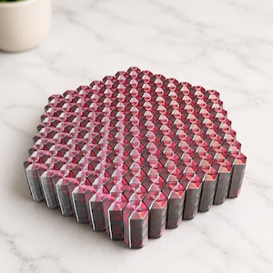 May include: A hexagonal trivet composed of numerous small, vertical, rectangular blocks arranged in a honeycomb pattern. Each block features a dark gray and magenta plaid pattern on its top surface, creating a textured, geometric grid design.