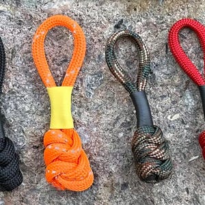 May include: Four colorful zipper pulls with knotted ends. The pulls are black, orange, camouflage, and red. Each pull has a loop at the top and a black or yellow accent. The pulls are on a textured gray surface.