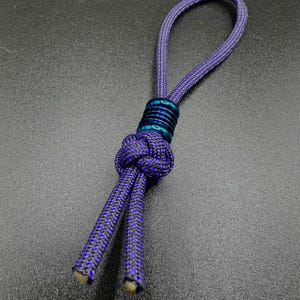 May include: A purple and black rope keychain with a loop and a decorative knot. The keychain features a blue metal accent piece. The rope is made of a durable material, suitable for everyday use.