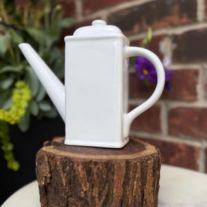 May include: A white ceramic watering can with a square body, a long spout, and a curved handle. The lid is small and rounded. The watering can is displayed on a wooden stump, with a brick wall and greenery in the background.