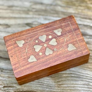 May include: A rectangular wooden box featuring a polished brown finish and brass inlay. The lid displays a central circular motif composed of four heart-shaped brass pieces, surrounded by additional heart-shaped accents and delicate engraved lines.