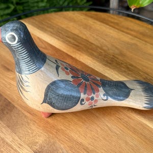 May include: Hand-painted ceramic bird figurine. The bird has a black head and tail, a beige body, and red and black floral designs. The beak is red, and the feet are pink. The figurine is on a wooden surface.
